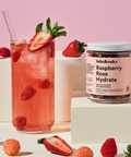 A glass of pink raspberry rose hydrate drink with strawberries and mint next to a jar of lake&oaks Raspberry Rose Hydrate, styled on white blocks with fresh raspberries against a pink background.
