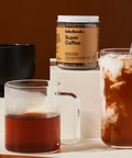 A glass jar labeled Lake & Onz Super Coffee sits on a white pedestal, styled with a glass mug of brewed coffee, a tall glass of iced coffee with milk, and a black mug against a warm brown background.