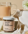 A jar of Lake & Oak 'Vanilla Cookie Calm' supplement tea blend in the foreground, styled with a glass mug of iced coffee, a honey dipper, and cookies on a neutral beige background.