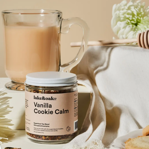 A jar of Lake & Oak 'Vanilla Cookie Calm' supplement tea blend in the foreground, styled with a glass mug of iced coffee, a honey dipper, and cookies on a neutral beige background.