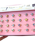 A hand holds an open pink jewelry box displaying 20 gold rings with colorful, iridescent rhinestone settings. The inner lid features branding text including 'Crazy Gemstones' and 'ORBUE'.