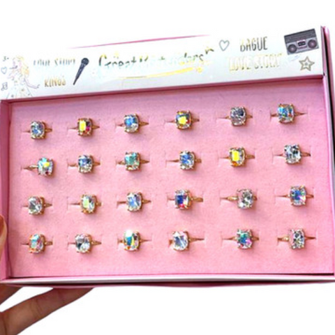 A hand holds an open pink jewelry box displaying 20 gold rings with colorful, iridescent rhinestone settings. The inner lid features branding text including 'Crazy Gemstones' and 'ORBUE'.
