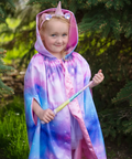 A young girl wearing a whimsical unicorn cape with a pink horn and ears, featuring a purple and blue galaxy tie-dye pattern, holding a wand in an outdoor garden setting.