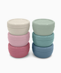 Two stacks of round, stackable storage containers with screw-on lids in pastel colors, photographed on a clean white background. The left stack contains cream, light pink, and dusty rose containers, while the right stack contains mint green, teal, and slate blue containers.