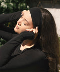 A woman outdoors wearing a black long-sleeve top, black headband, and black fingerless gloves, posing with her eyes closed.