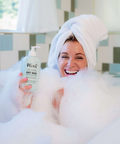 Woman in a bathtub with bubbles holding a bottle of body wash, wearing a towel on her head.