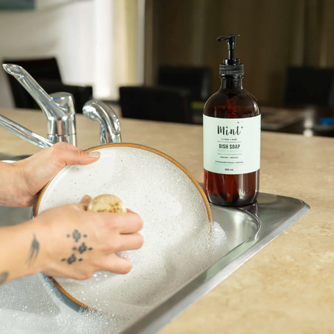 Person washing a dish in a sink with a bottle of dish soap on a counter.