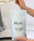 A person holding a bag of Mint brand fabric softener with a spoonful of the white powder being showcased in front of a washing machine.