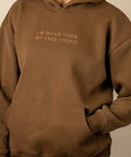A person wearing a brown pullover hoodie with the text 'I'M NICER THAN MY FACE LOOKS' printed in gold on the chest, paired with light gray jeans against a plain beige background.