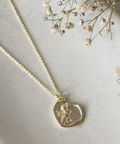 A gold chain necklace with a small, irregularly shaped gold pendant featuring an engraved figure, styled on a textured light beige ceramic surface with dried baby's breath flowers.
