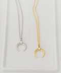 Two delicate crescent moon necklaces on a white tray, one in silver and one in yellow gold, with their chains extending out of frame.