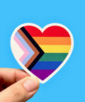 Heart-shaped sticker with rainbow and plus symbol held by a hand against a blue background