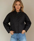 Woman wearing a black quilted jacket and blue jeans against a beige background