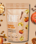 A stand-up pouch of Eatable Pumpkin Pop Toddly gourmet popcorn, surrounded by decorative autumn ingredients including apples, cinnamon sticks, star anise, dried orange slices, and more popcorn on a warm beige background.