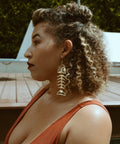 A model sits in profile by a pool while wearing gold mirror acrylic Medium Salmon Ghost earrings from Copper Canoe Woman.