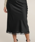 A black midi skirt with a sheer lace hem, worn by a model styled with a beige polka dot cardigan and black heels against a light beige background.
