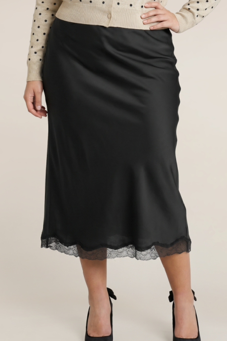 A black midi skirt with a sheer lace hem, worn by a model styled with a beige polka dot cardigan and black heels against a light beige background.