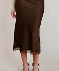 A woman wearing a brown satin midi skirt with a lace hem, styled with a cream polka-dot cardigan and black pointed-toe heels against a light gray studio background.