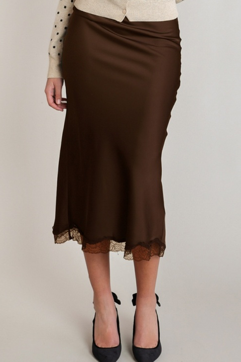 A woman wearing a brown satin midi skirt with a lace hem, styled with a cream polka-dot cardigan and black pointed-toe heels against a light gray studio background.