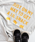 White t-shirt with yellow text and fish graphics, paired with black sneakers on a white background.