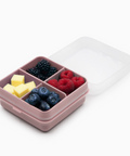 A pink compartment bento box with a clear lid, shown open and filled with blueberries, blackberries, raspberries, and cheese cubes on a white background.