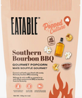A package of Eatable Popped Culture Southern Bourbon BBQ Gourmet Popcorn on a clean white background. The packaging is a warm peach-beige color with orange accents, featuring illustrations of a bottle of oil, popcorn kernels, and a small bowl of dark seasoning.