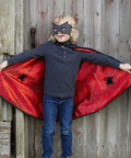 A young child wearing a black spider-themed mask and a red cape with black spiderweb patterns and spider graphics, standing in front of a weathered wooden plank wall.