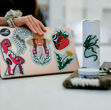 Person holding a laptop with stickers, surrounded by various items on a table.