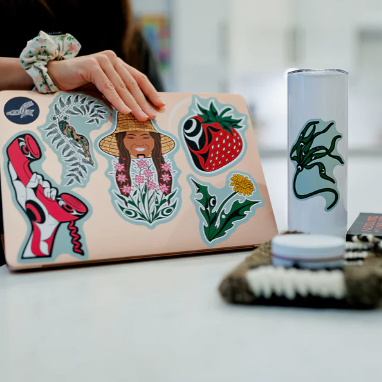 Person holding a laptop with stickers, surrounded by various items on a desk.
