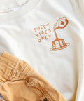 White t-shirt with 'Sweet Vibes Only' text and graphic, paired with beige shorts.