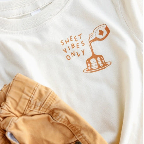 White t-shirt with 'Sweet Vibes Only' text and graphic, paired with beige shorts.