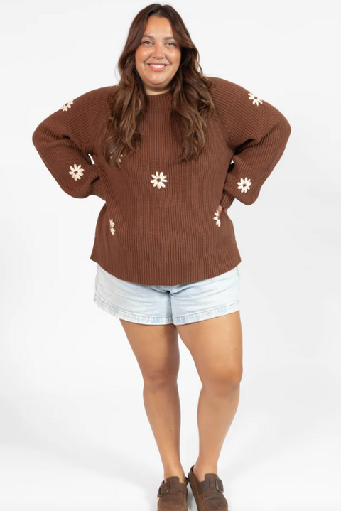 Person wearing a brown sweater with white floral patterns and light blue shorts on a white background