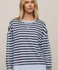 A woman wearing a navy and light gray striped long-sleeve knit sweater with a ribbed hem and cuffs, paired with light blue shorts, against a solid beige background.