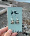 Pair of earrings on a 'Tide and Timber' branded card held over a beach background