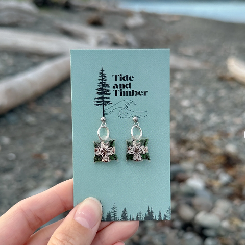 Pair of earrings on a 'Tide and Timber' branded card held over a beach background