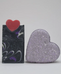 Two heart-shaped soap bars, one black and white marbled and the other purple glittery, on a light gray background.