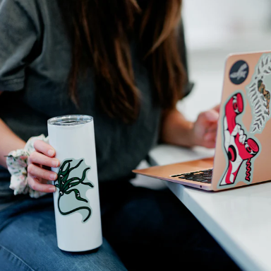 Person using a laptop with stickers on a desk, holding a tumbler.