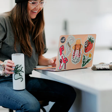Person sitting at a table with a laptop, holding a tumbler, and surrounded by various items.