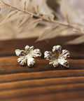 Gold flower-shaped earrings on a wooden surface with dried leaves in the background