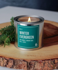 Candle in a metal can labeled 'Winter Evergreen' on a wooden surface with greenery.