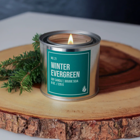 Candle in a metal can labeled 'Winter Evergreen' on a wooden surface with greenery.