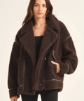 A woman modeling a dark brown sherpa fleece jacket with a wide collar and front pockets, paired with a black top and black pants, against a plain off-white background.
